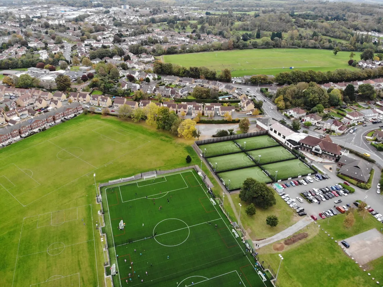 Aerial view of a sports complex with football pitches and tennis courts surrounded by suburban housing and green fields.