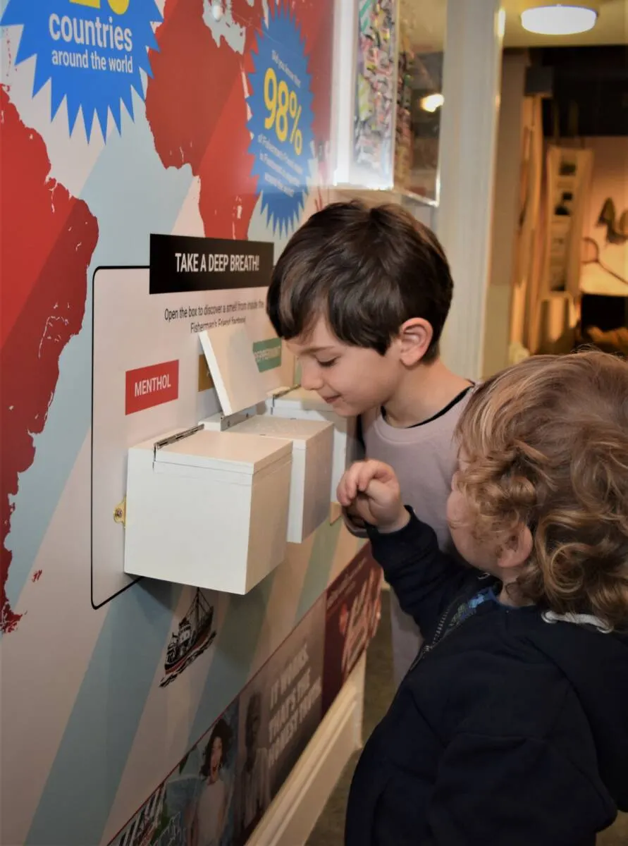 Two young children interact with an interactive museum display featuring a world map and a "Take a Deep Breath" scent-discovery box.