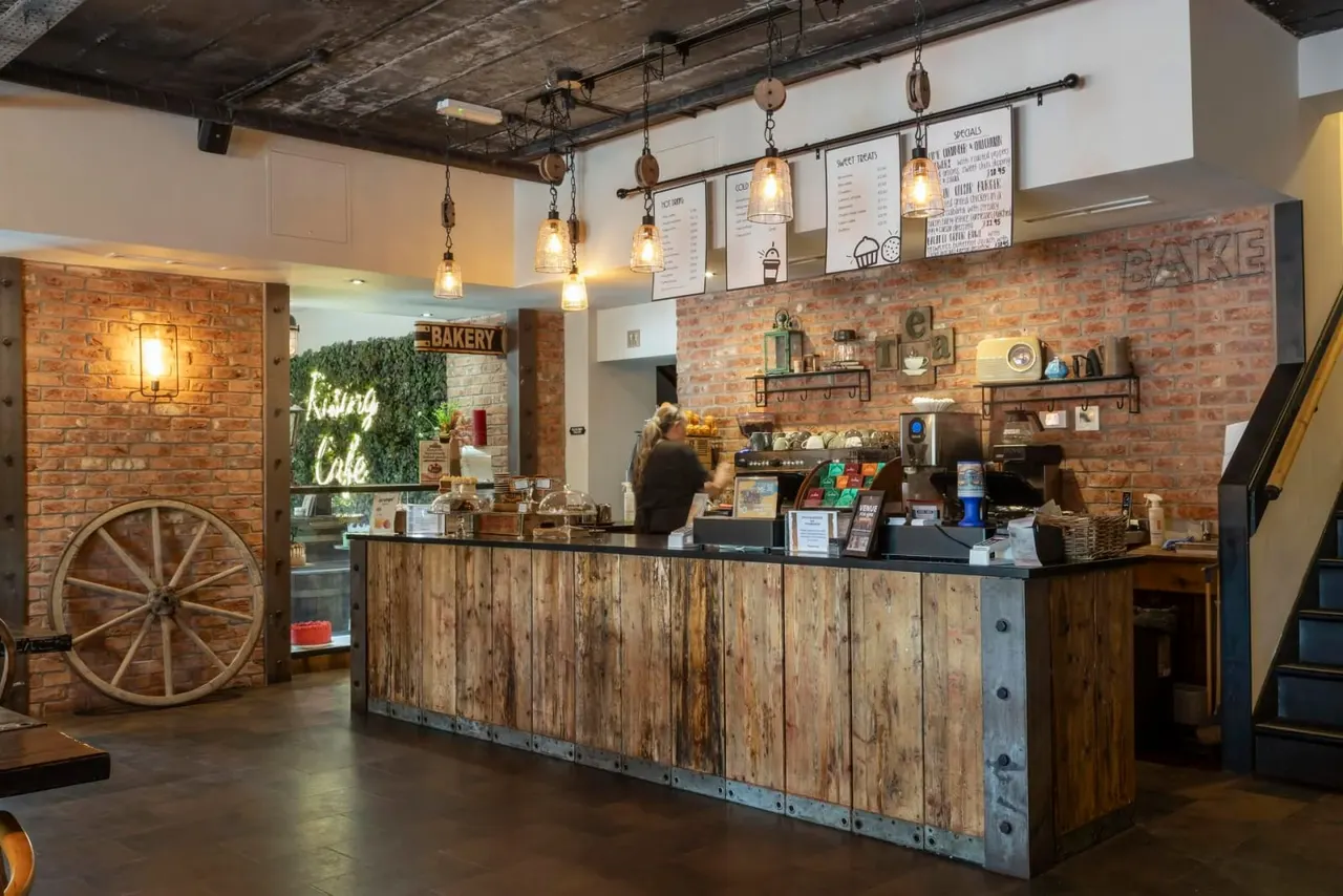 Interior of a rustic bakery café with exposed brick walls, vintage wooden counter, pendant lighting, and staff member behind the service desk.
