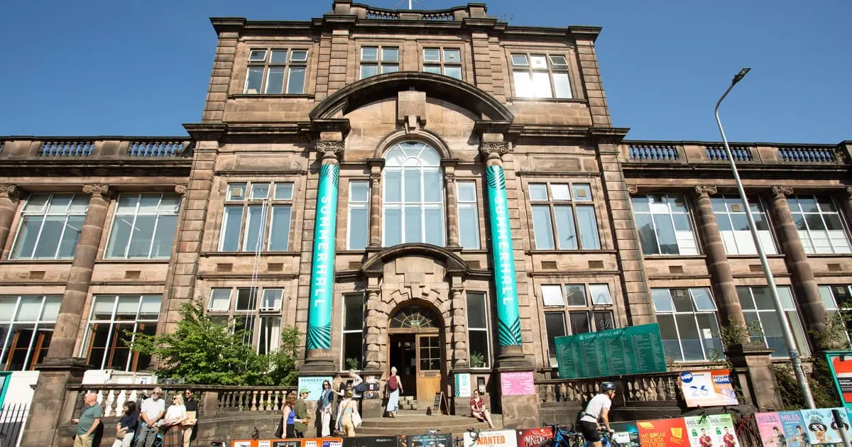 Historic stone building with turquoise banners, wide entrance steps, and visitors gathered outside on a sunny day.