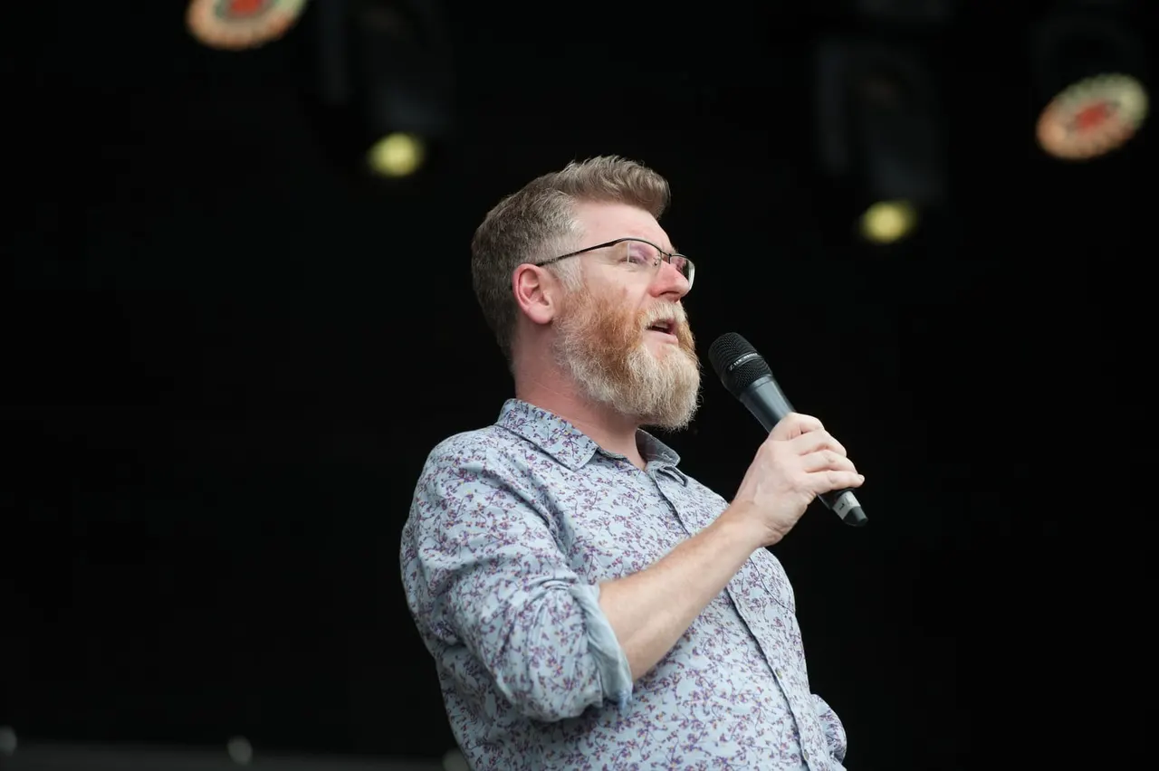 A man wearing glasses and a floral shirt speaks into a microphone on stage against a dark background.