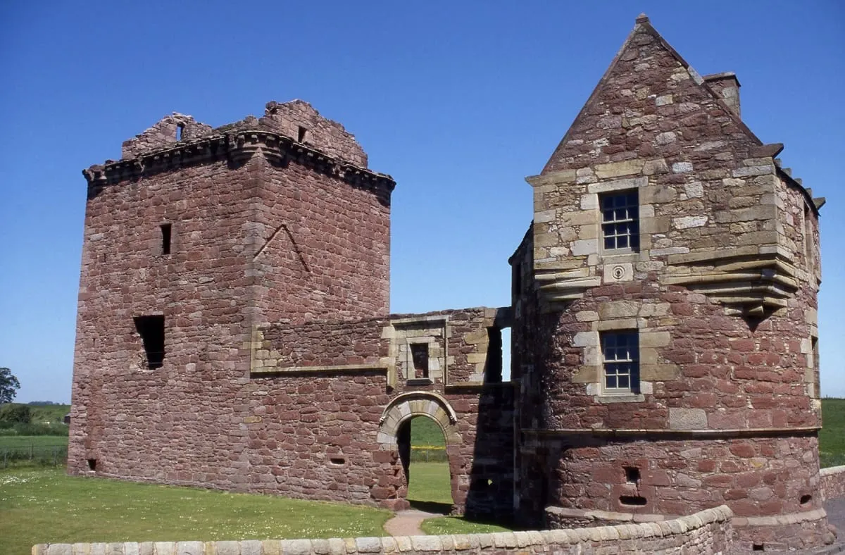 Ruined red sandstone castle with two towers and connecting walls set in grassland under clear blue sky.