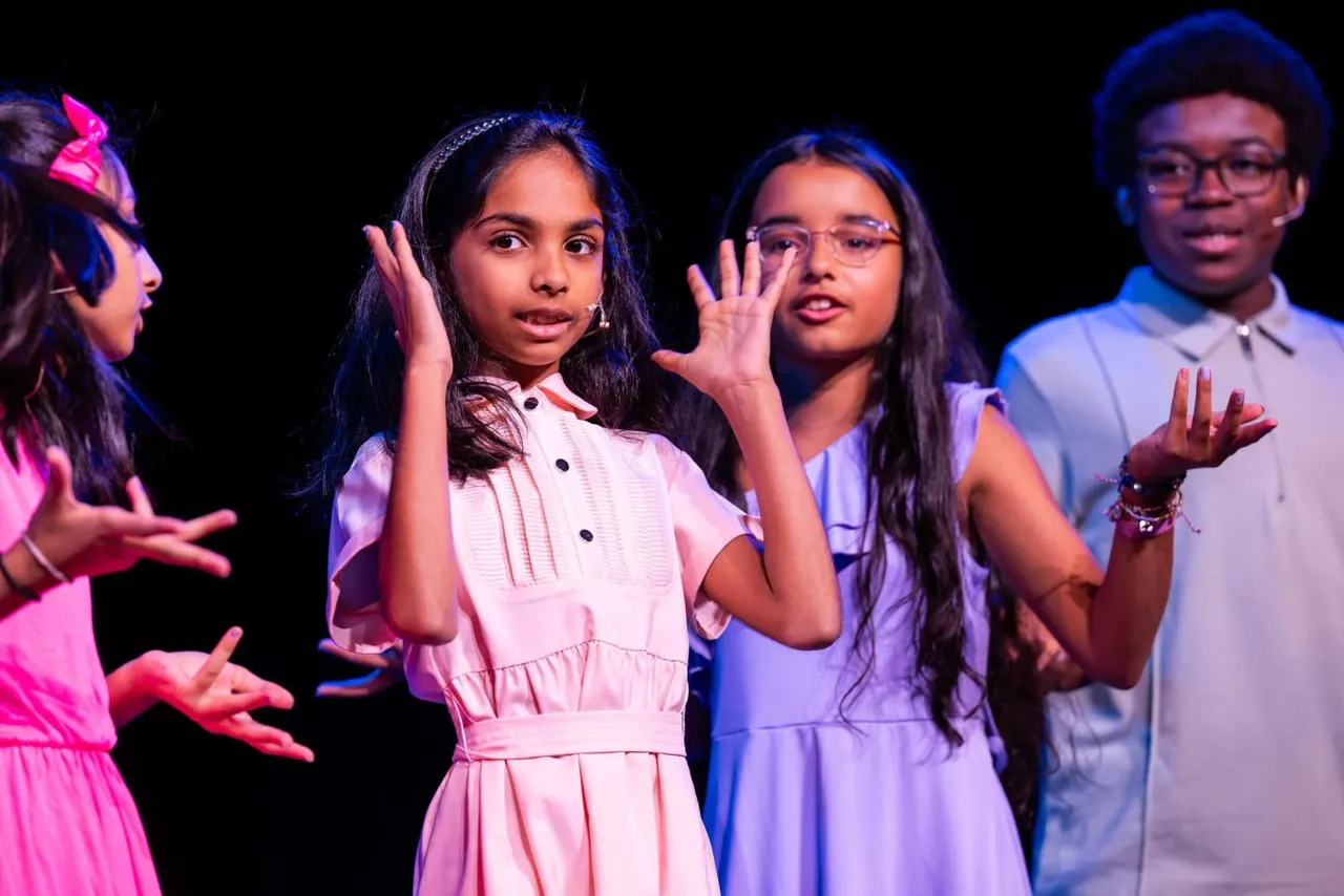 Young performers singing and gesturing expressively on a stage with coloured lighting.