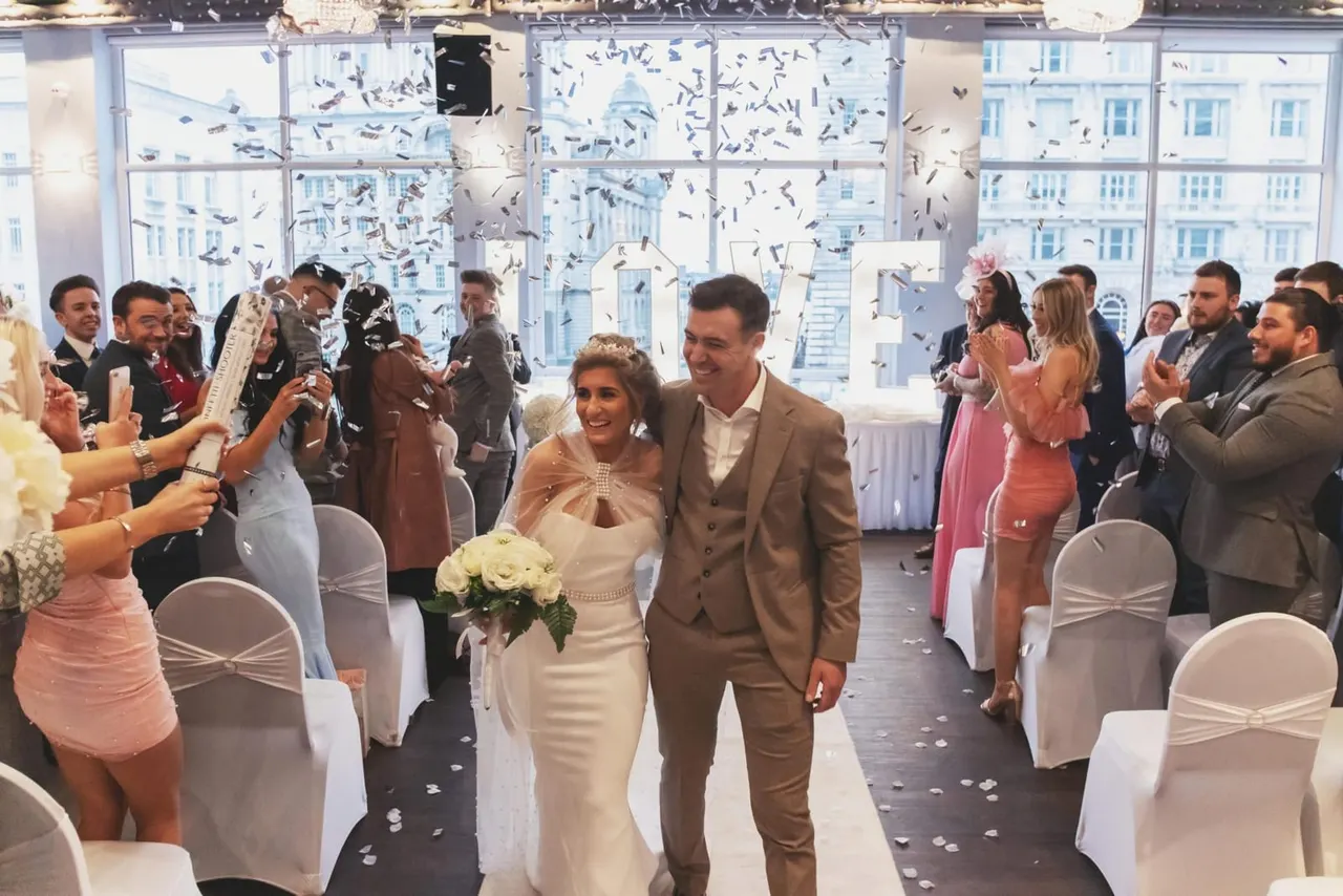 Bride and groom walking through confetti-filled aisle with guests celebrating at a wedding reception with city views.