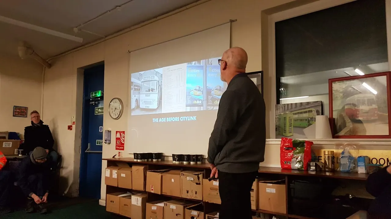 Man giving a presentation about CityLink to an audience in a bus garage, with a projector screen displaying historical transport imagery.