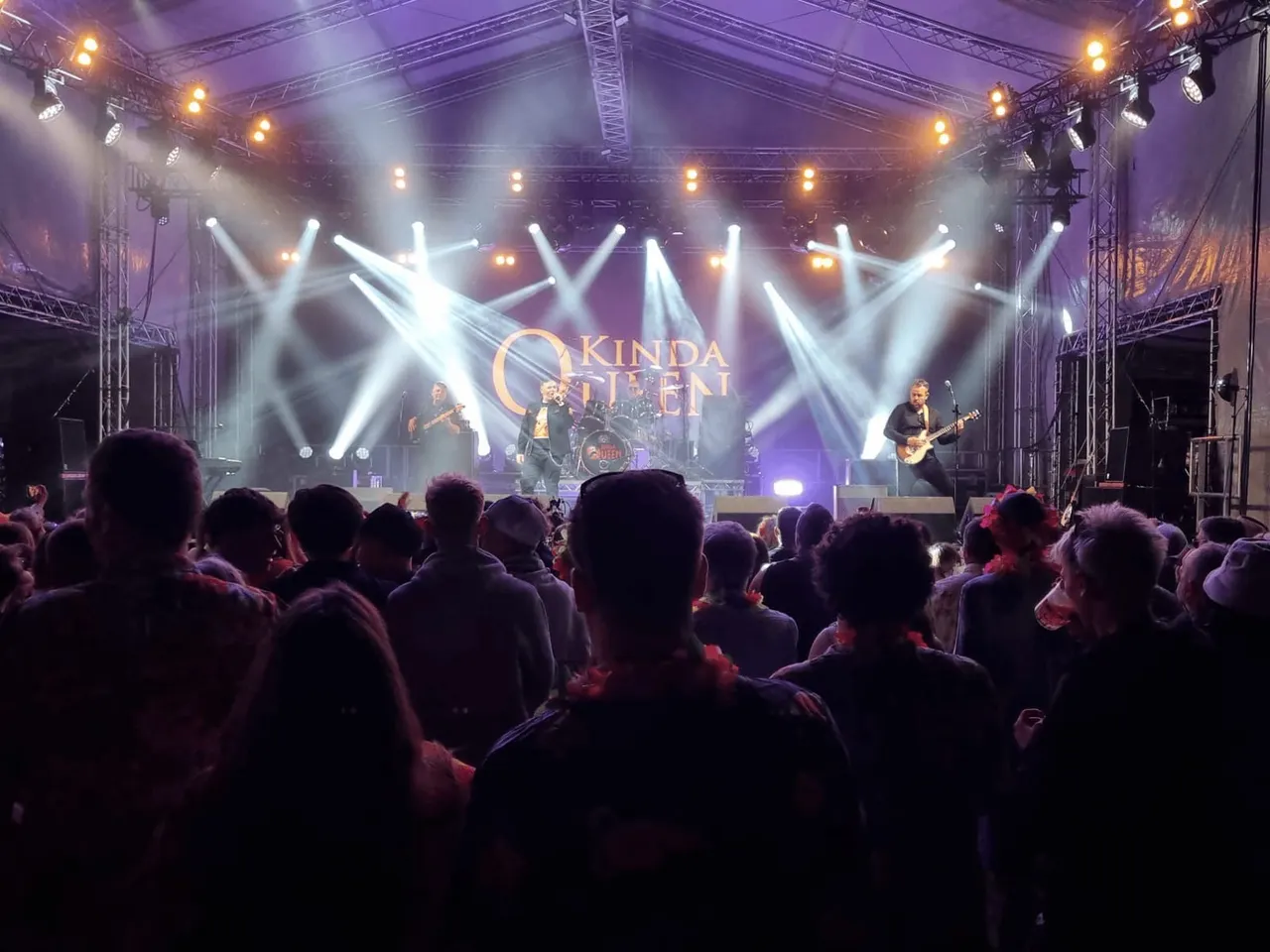 Live band performing on stage with purple lighting and dramatic spotlights whilst large crowd watches from the floor.