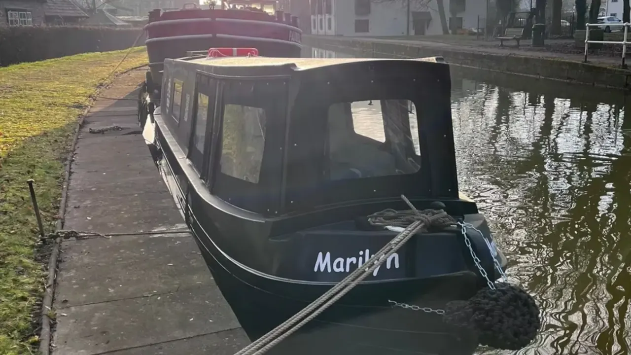 Black narrowboat moored alongside a canal towpath with residential buildings in the background.