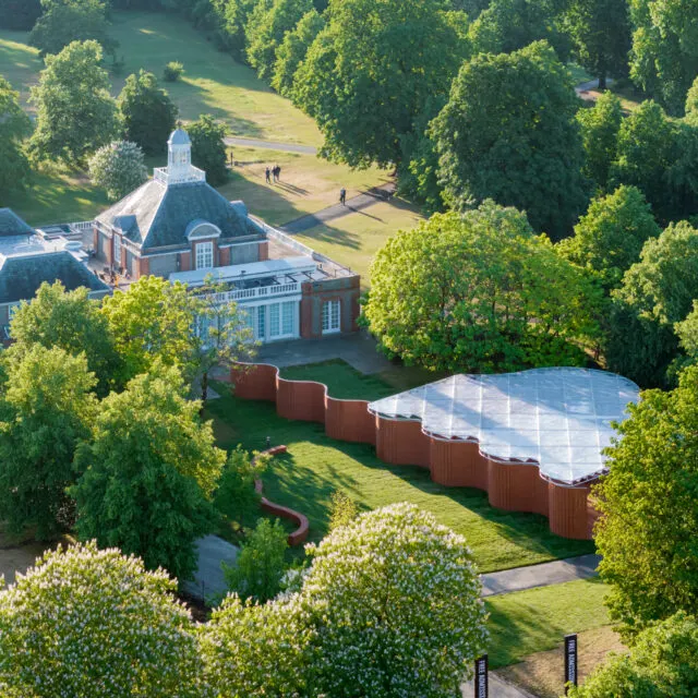 Aerial view of a brick building with a domed roof surrounded by mature trees and manicured gardens, with a white marquee tent alongside.