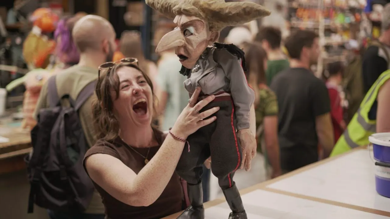 A delighted visitor holds a large bird puppet with an exaggerated beak while surrounded by other guests at an indoor event.