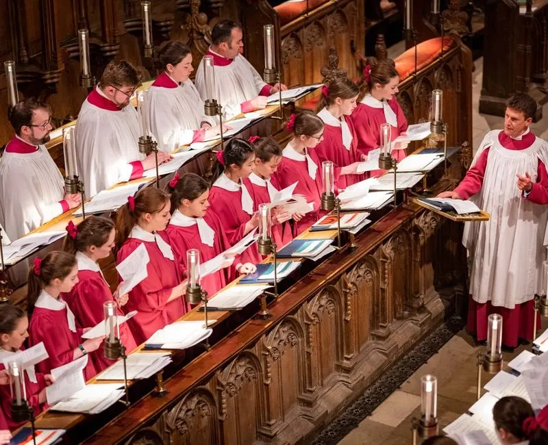 Cathedral choir members in red and white robes singing from ornate wooden stalls during a service.