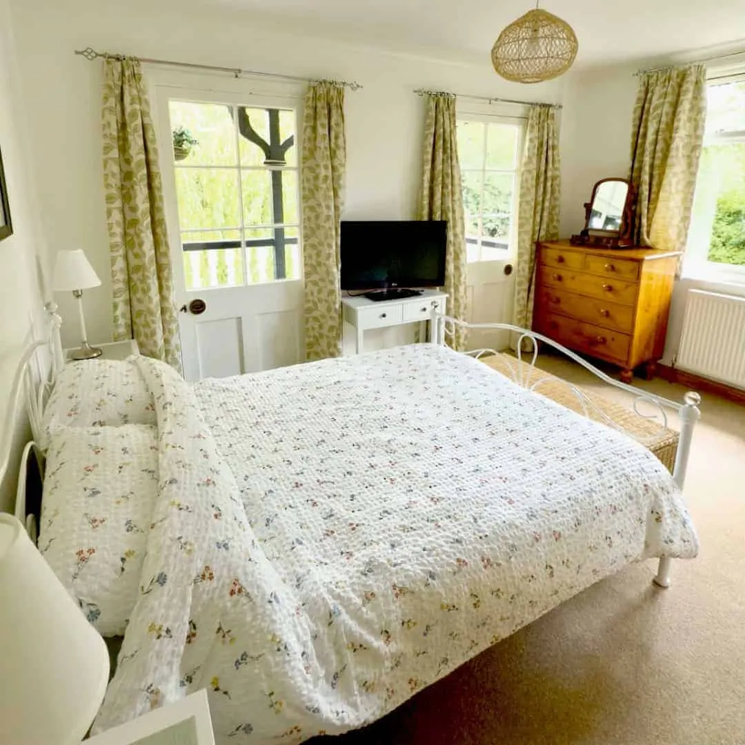 Bright bedroom with double bed, white metal bedframe, wooden chest of drawers, TV, and multiple windows with green patterned curtains overlooking gardens.