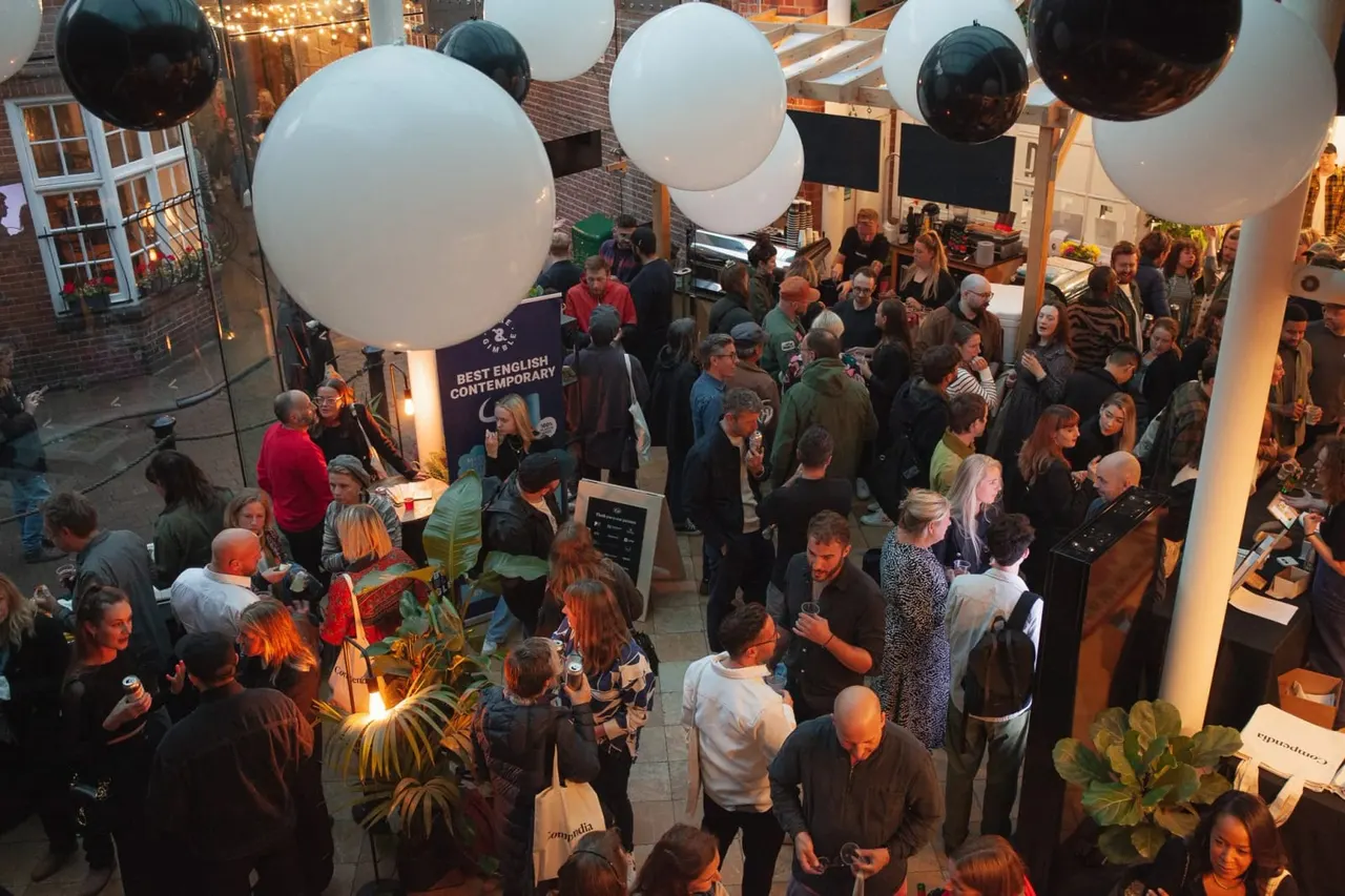 Overhead view of a crowded evening event in a brick courtyard with dozens of guests mingling under large illuminated white and black spherical balloons.