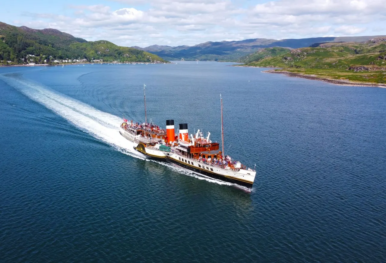 A vintage paddle steamer with passengers on deck cruises through a Scottish loch surrounded by green hills and mountains.