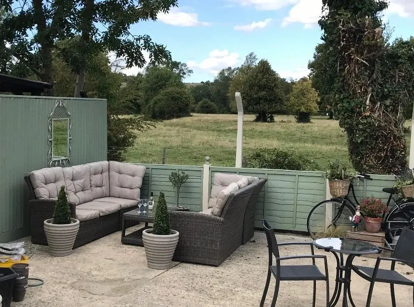 Outdoor seating area with rattan furniture, potted plants and a bicycle, overlooking open fields and woodland.