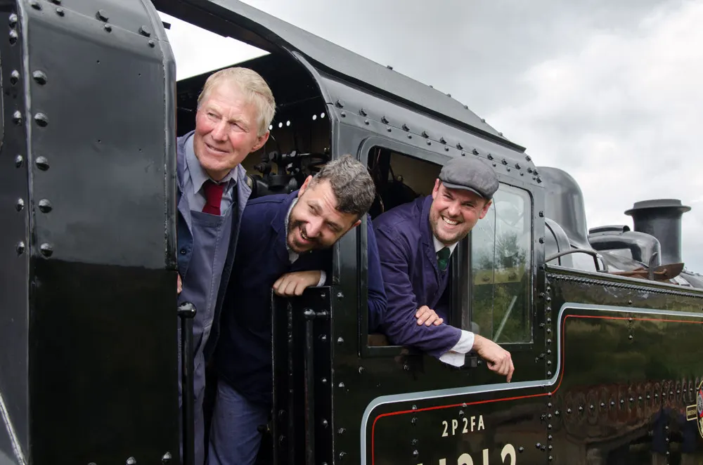 Three men in period uniforms leaning from the cab of a heritage steam locomotive.