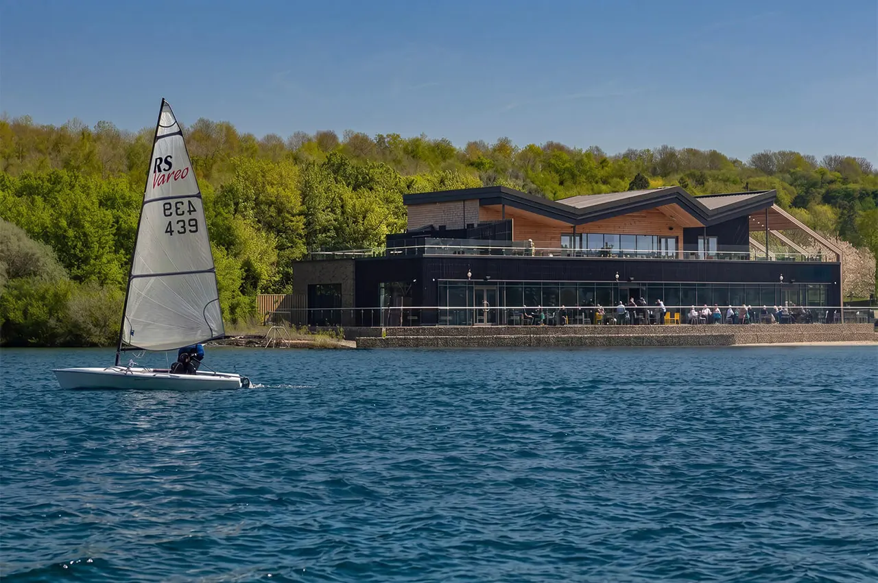 Sailor in a small white dinghy with sail, on water in front of a modern clubhouse with spectators on a terrace.