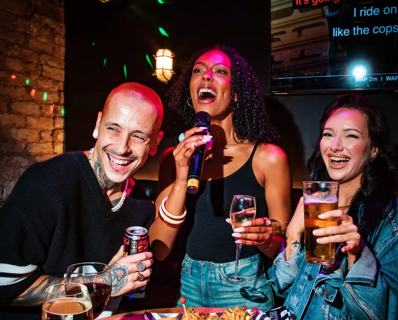 Four people singing karaoke with drinks at a lively nightclub venue illuminated by pink and green stage lighting.
