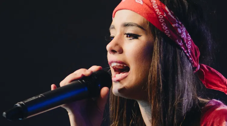 Woman in red headwrap singing into microphone against black background.