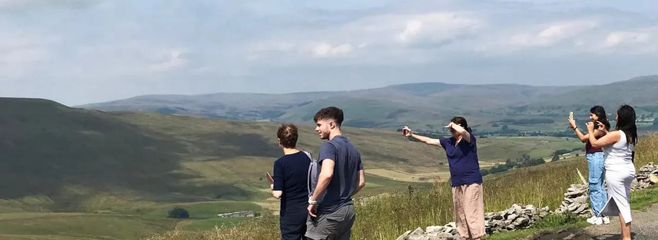 A group of visitors stand on a hillside overlooking expansive moorland and rolling valleys under a cloudy sky.