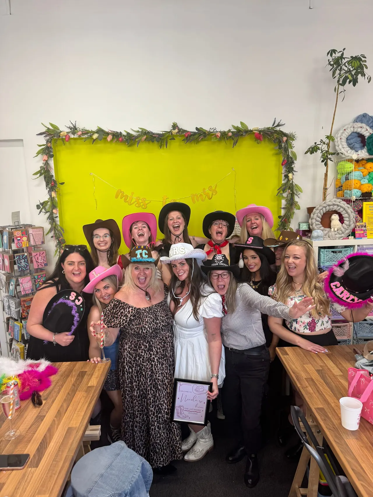 Large group of women posing together in a brightly decorated craft space with a neon yellow backdrop and floral garland, wearing colourful hats and festive attire.