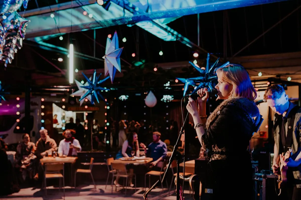 A woman singing at a microphone in a festively decorated indoor venue with hanging stars and colourful lights, surrounded by seated guests.