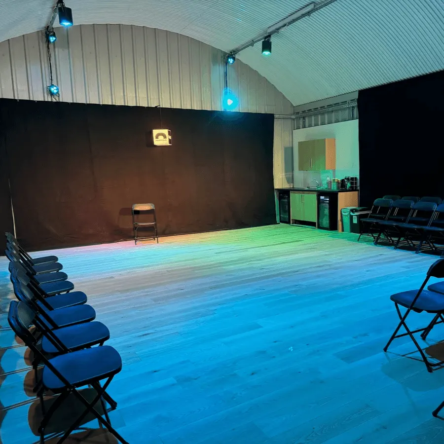 A theatre studio with a lit stage, black backdrop, blue lighting on the floor, and rows of folding chairs arranged for an audience.