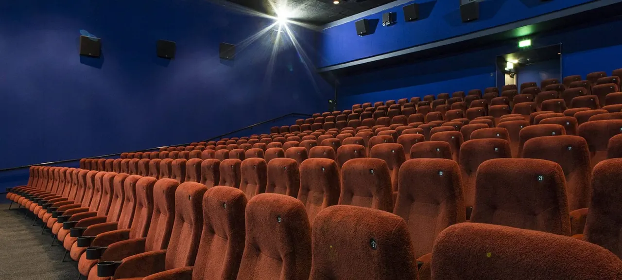 Empty cinema auditorium with rows of red seats facing the screen, blue walls and lighting.