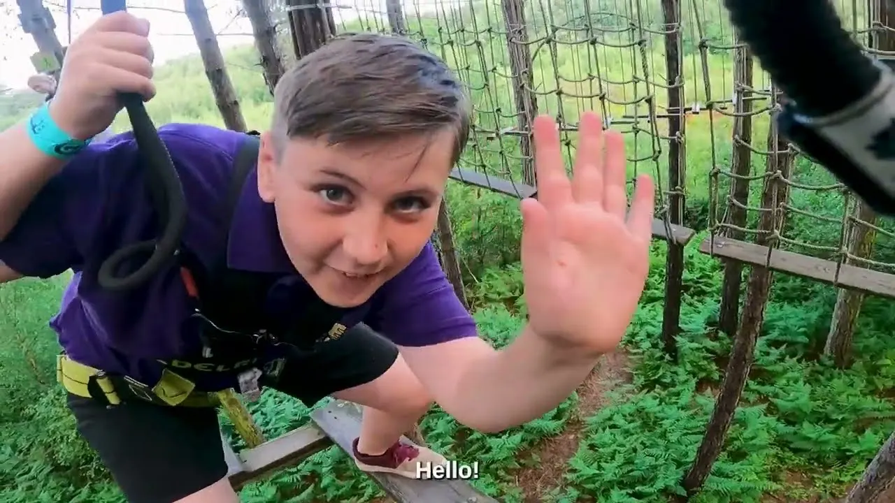Child on a zip-wire course among tree structures at an outdoor adventure park, waving at the camera.