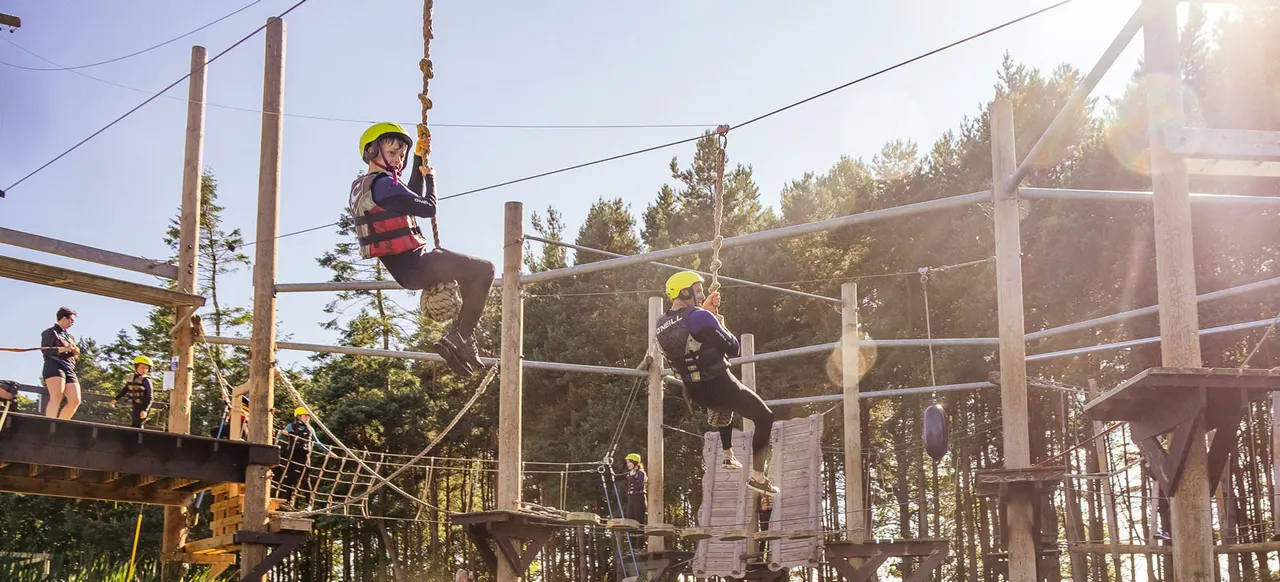 People wearing helmets navigate a rope course with wooden structures and zip lines amongst forest trees.