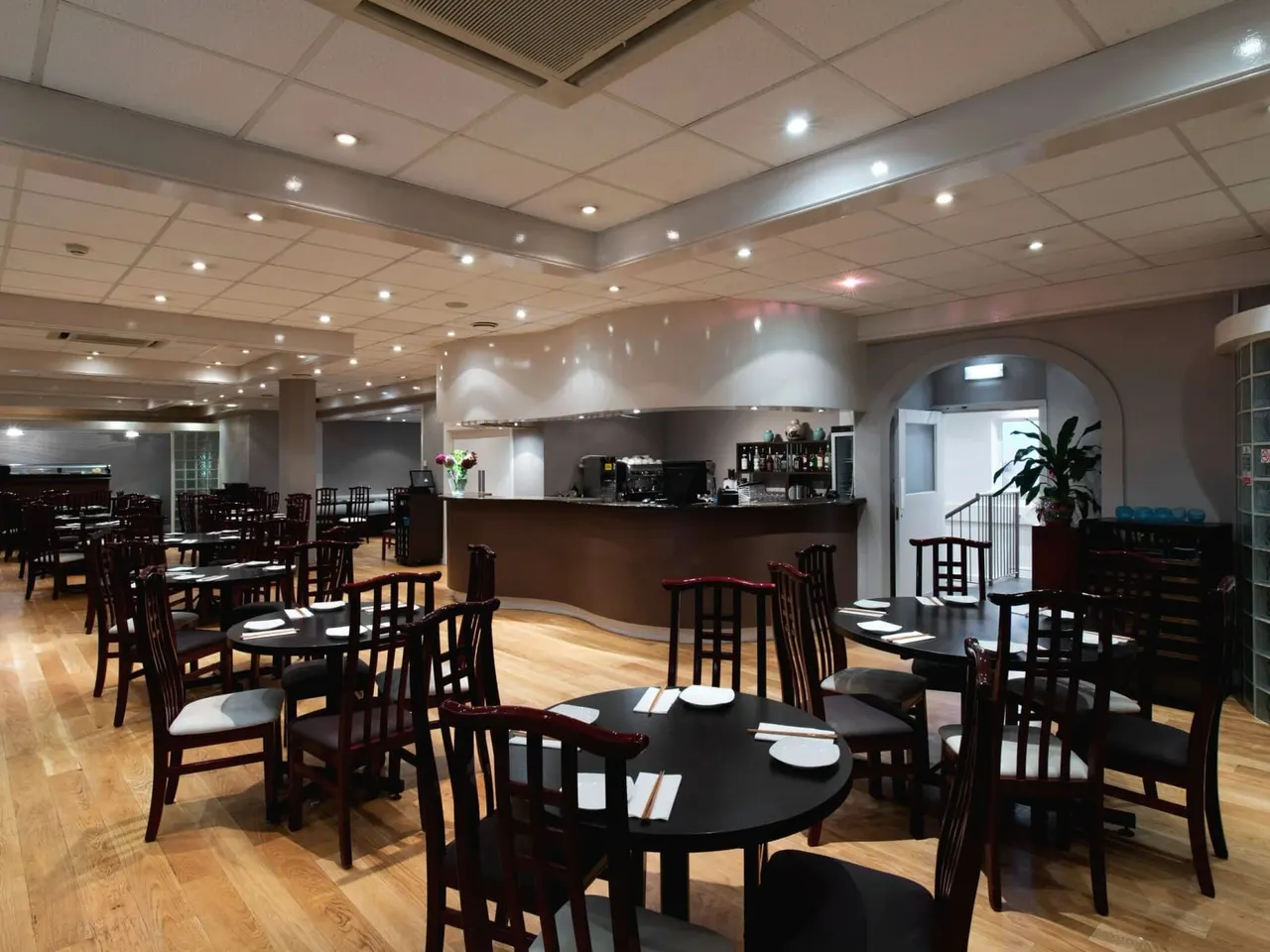 Interior of a modern restaurant dining room with dark wood furniture, round black tables set with place settings, and a service counter in the background.