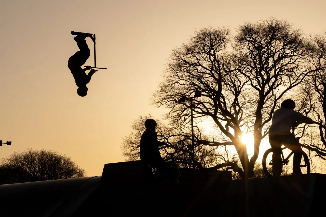 Silhouetted figures performing tricks at a skatepark at sunset, with one person airborne on a skateboard and others on bikes.