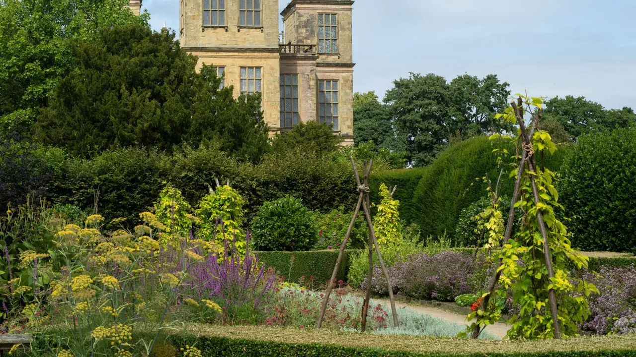 A formal walled garden with climbing plants on wooden supports, herbaceous borders and hedges, with a stone country house tower visible behind.