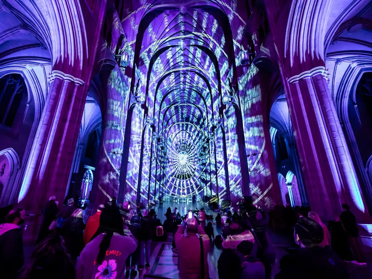 Interior of Norwich Cathedral illuminated with purple and blue lighting, with visitors walking through the nave beneath Gothic arches.