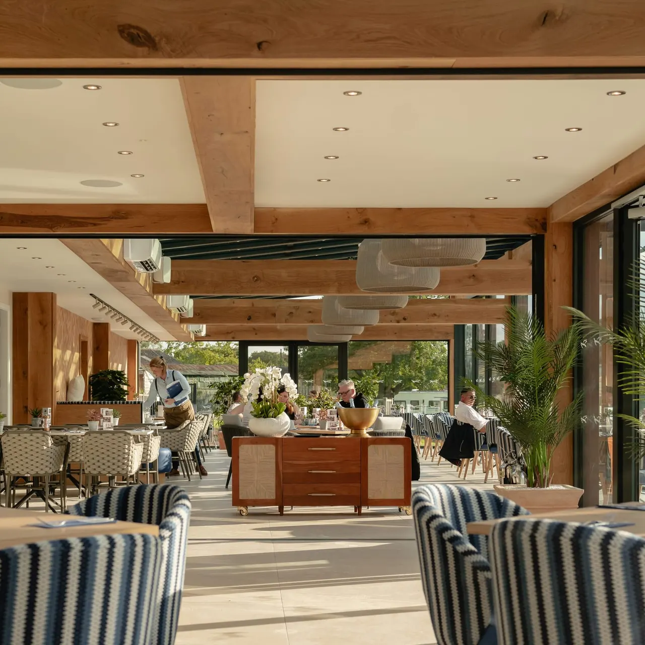 Modern indoor-outdoor dining space with wooden beams, striped armchairs, and guests seated at tables with staff attending.