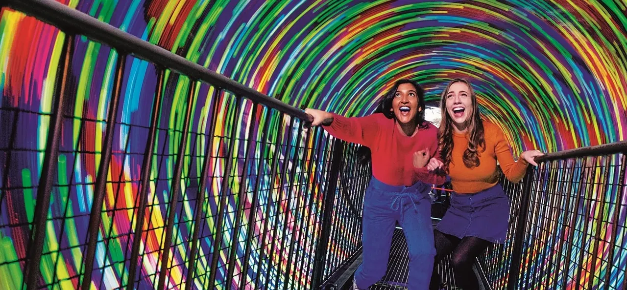 Two visitors walking through a vibrant rainbow-coloured light tunnel installation with animated expressions.