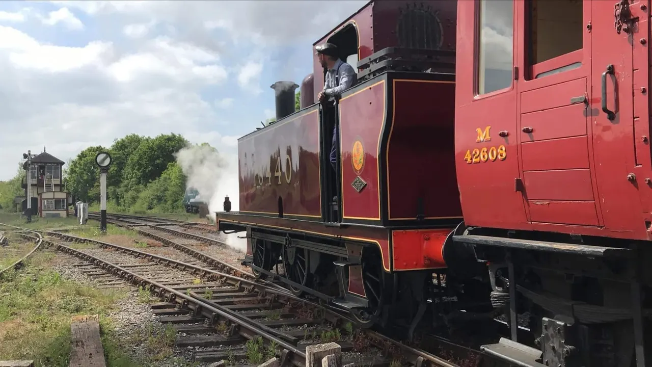 A fireman stands on a heritage steam locomotive at a railway station, with another steam engine visible in the distance emitting smoke.