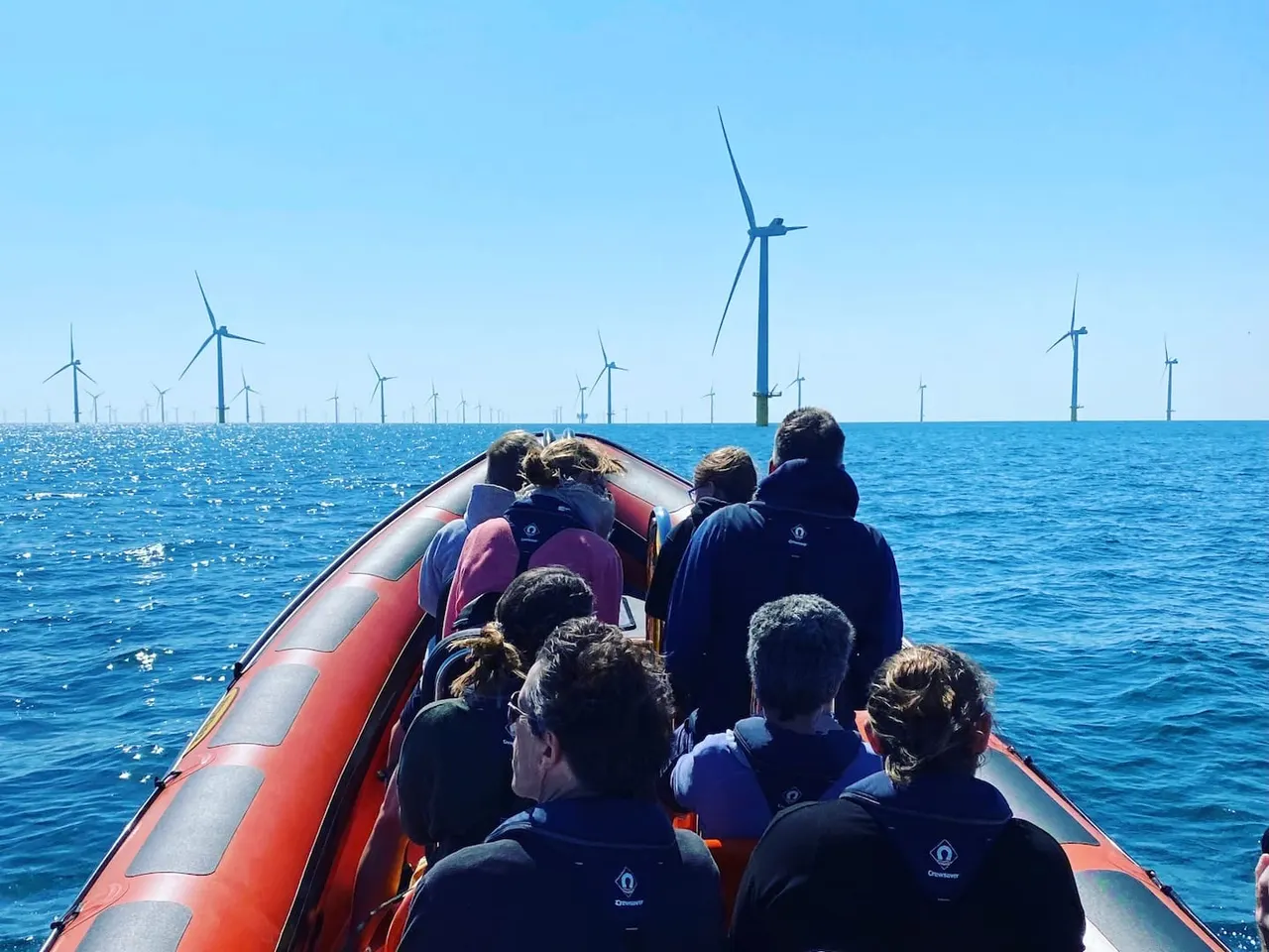 Passengers in a red speedboat heading toward offshore wind turbines in clear blue sea.