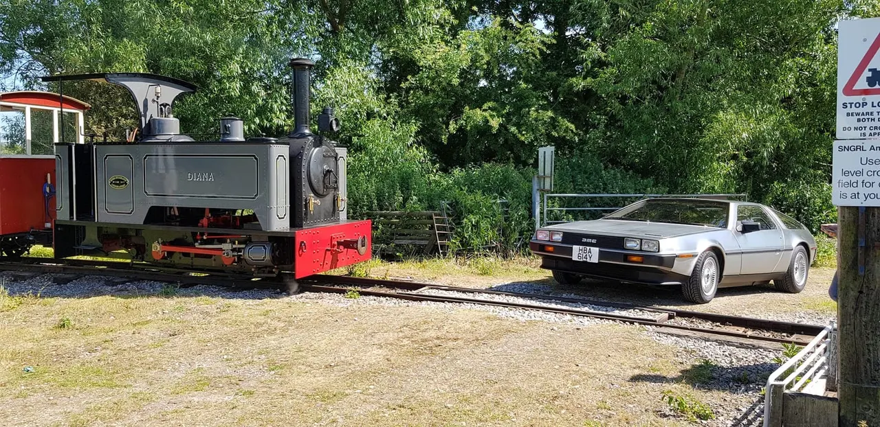 A heritage steam locomotive named Diana alongside a vintage sports car on a narrow-gauge railway track.