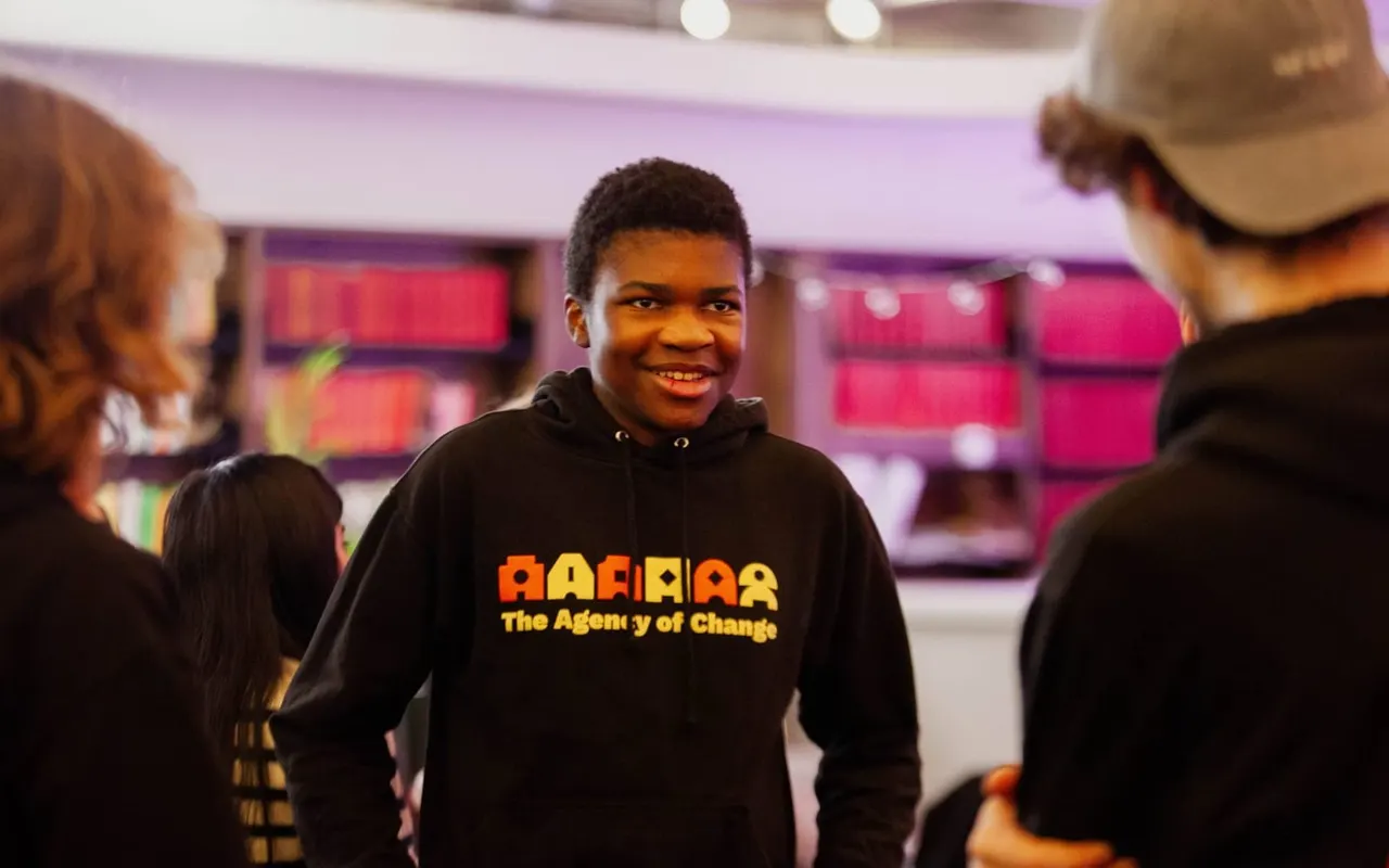 Young person smiling at camera surrounded by others in an indoor venue with pink and purple lighting.