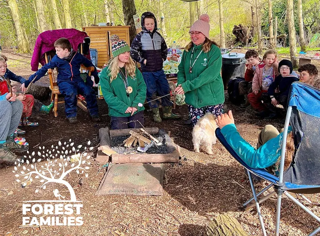 Children and adults gathered around a campfire in a woodland clearing during a supervised outdoor activity session.