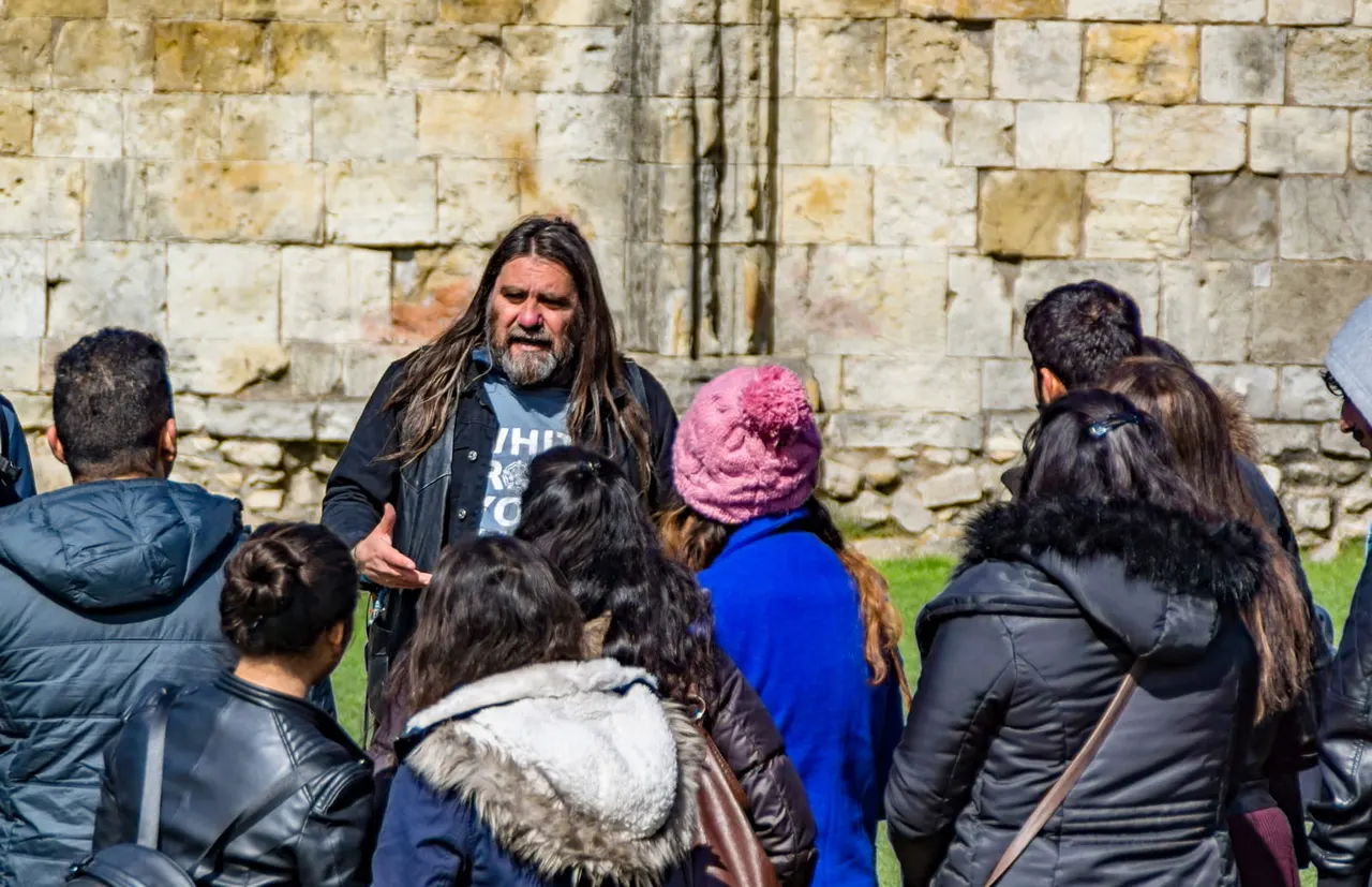 A guide with long hair and beard addresses a group of visitors gathered in front of a historic stone wall.