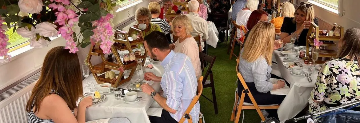 Groups of people seated at white-clothed tables enjoying a meal in a bright indoor venue decorated with pink flowers and artificial grass.