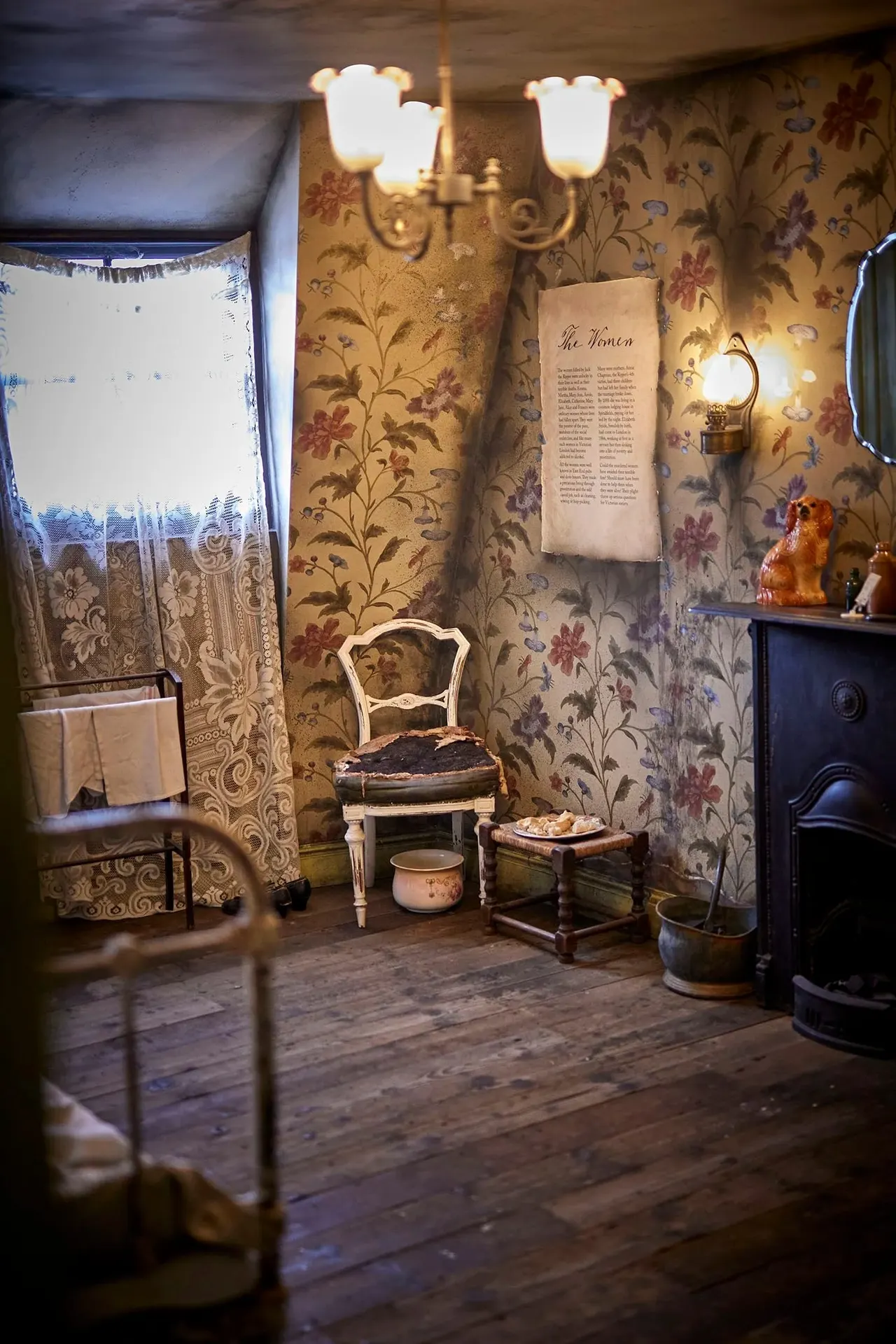 Period bedroom display with floral wallpaper, lit by wall sconces and a chandelier, furnished with antique chair, small table, and historical artefacts.