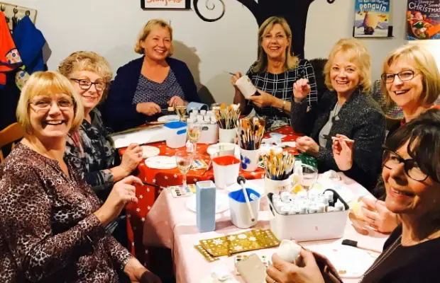 Group of smiling women seated around a table at a pottery painting class, holding brushes.