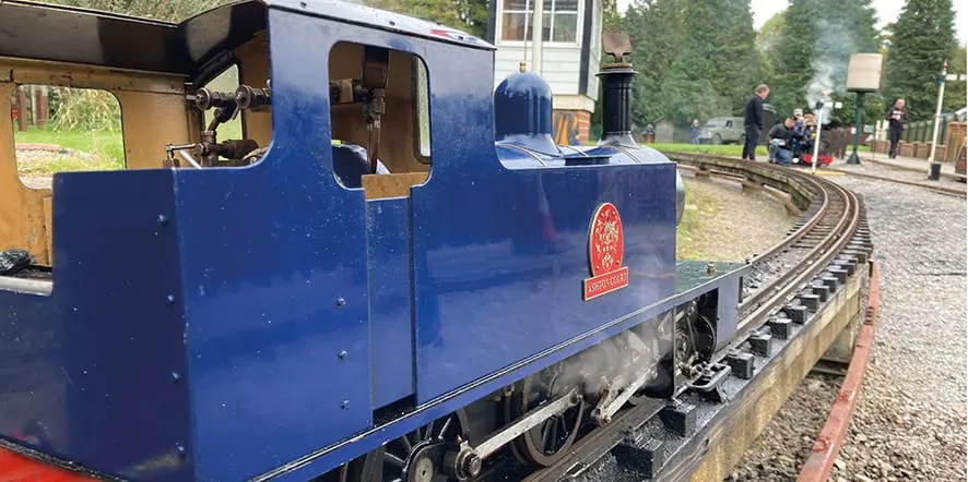 A blue miniature steam locomotive on narrow-gauge track at a railway attraction, with visitors in the background.