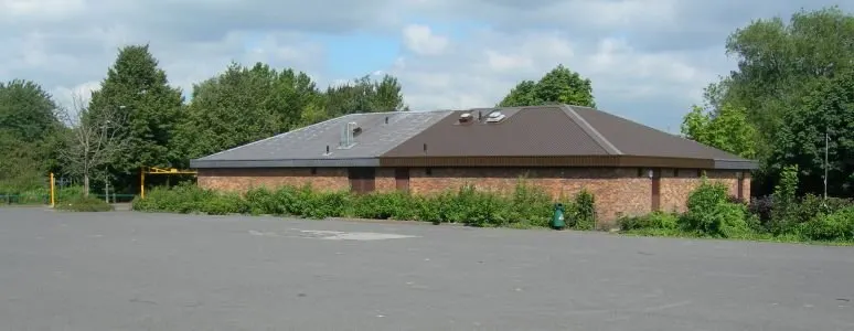A single-storey brick building with a dark roof set in a car park, surrounded by trees.