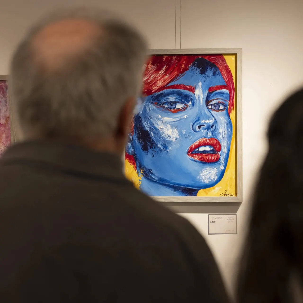 Two visitors viewing a bold pop art portrait with blue face, red hair and lips, and yellow background in a gallery setting.