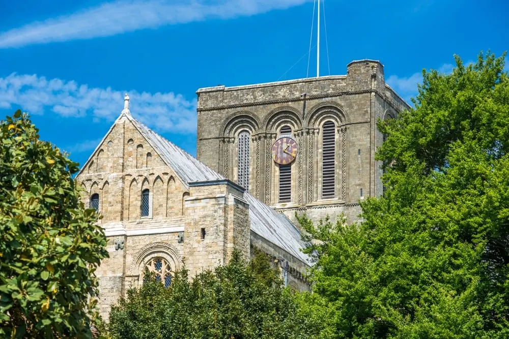 Historic stone church with clock tower surrounded by green foliage under blue sky.