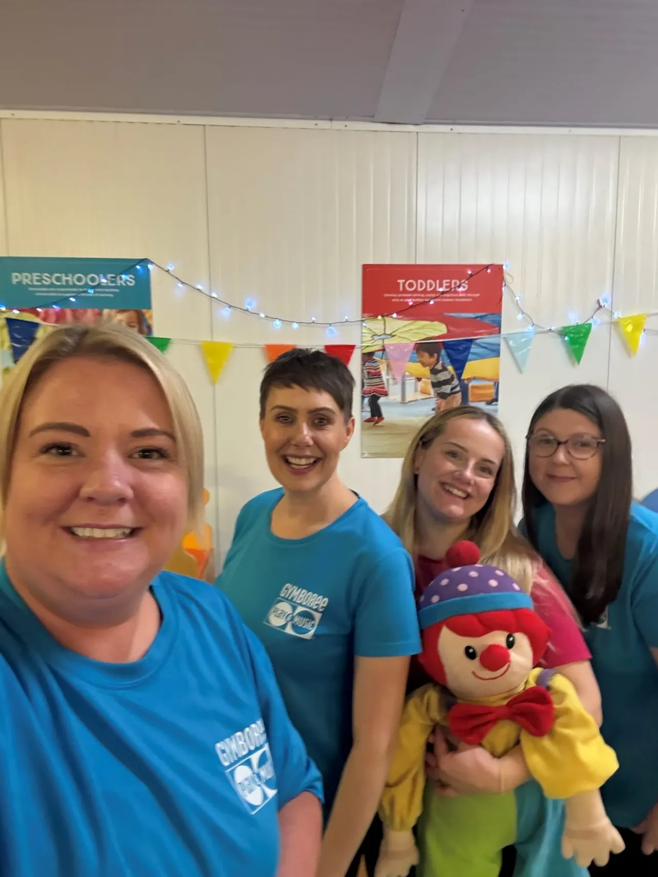 Four smiling staff members in blue uniforms pose together holding a colourful clown toy in a decorated playroom.