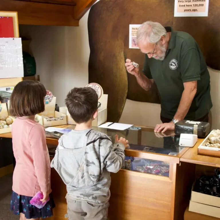 An older man in a green polo shirt speaks to two young children at a museum display counter with fossils and artefacts.